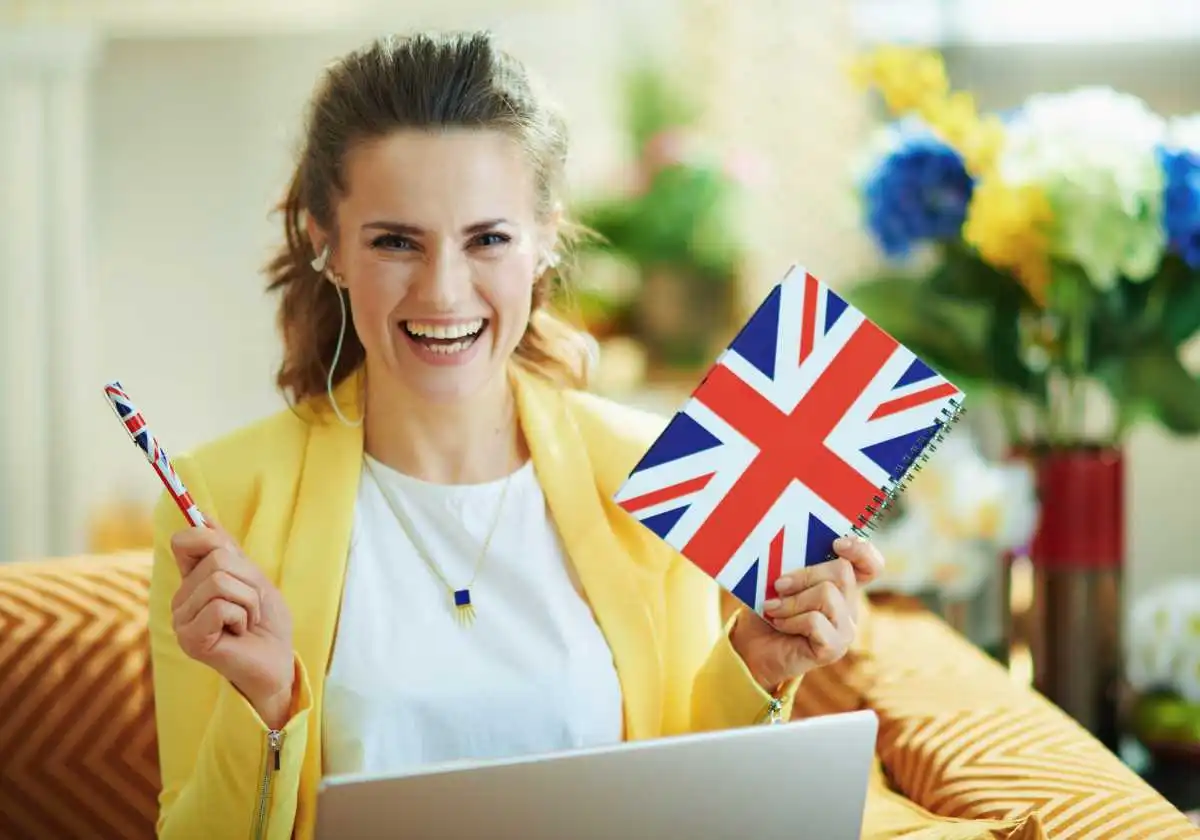 Smiling girl student having Bachelor Degree in UK holding a pen and UK flag and working on laptop